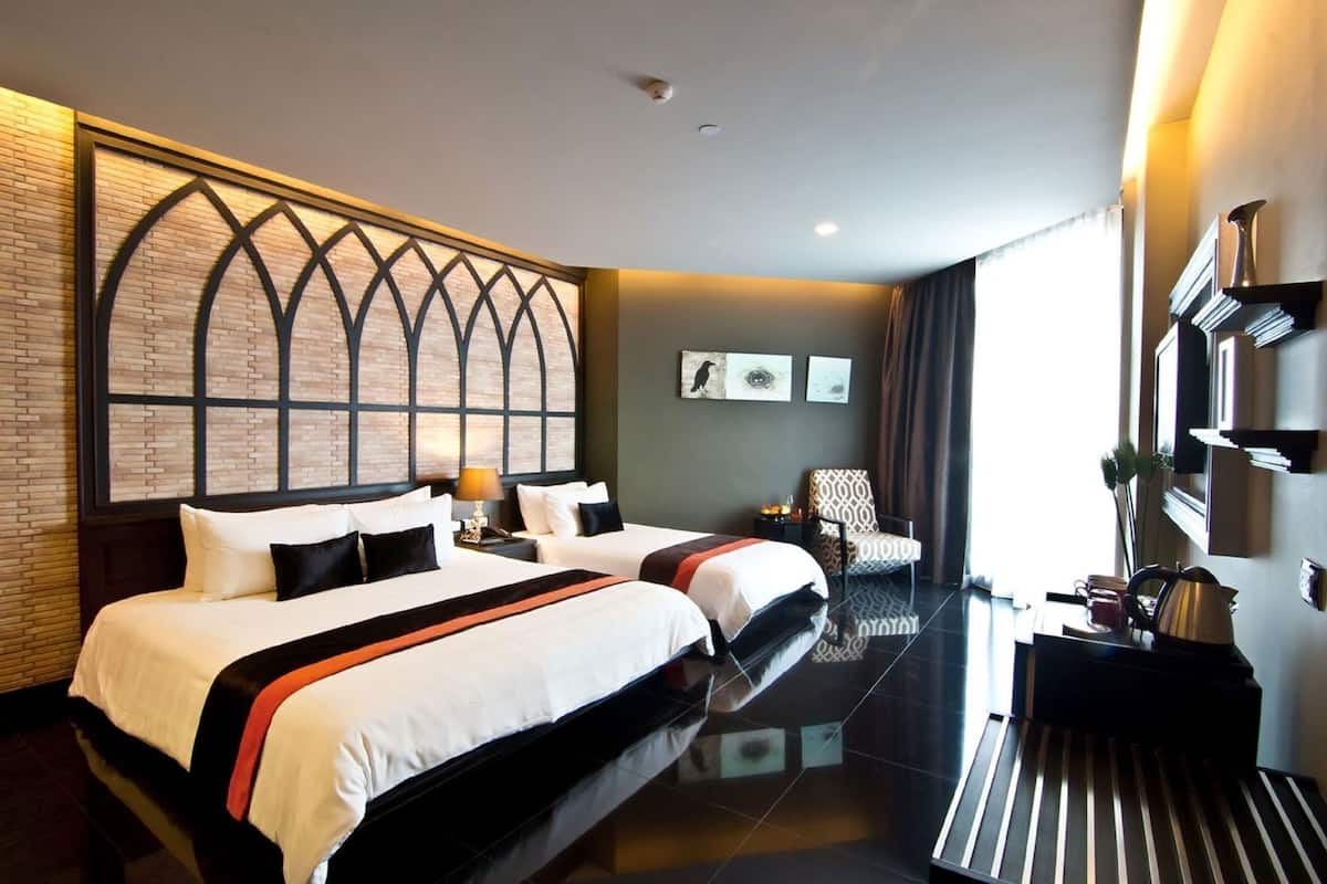 Quarto de hotel com duas camas, uma escrivaninha, uma cadeira, uma janela grande e um painel decorativo na parede.