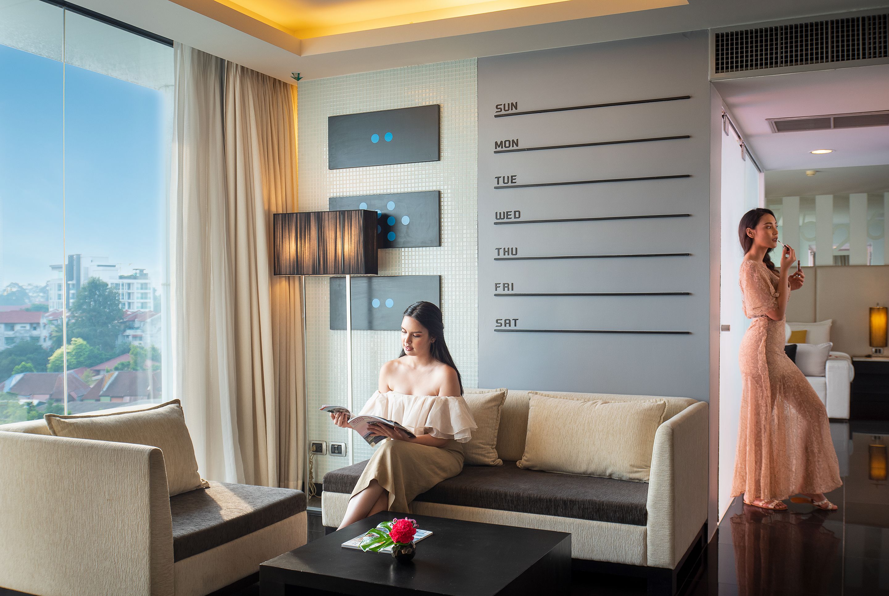 A modern hotel room with a large window, a sofa, a coffee table with a vase, and a wall calendar.