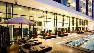 A modern hotel lobby with a pool area, featuring sun loungers, umbrellas, and a glass facade.