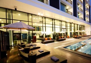 Un hall d’hôtel moderne avec un espace piscine, comprenant des chaises longues, des parasols et une façade en verre.