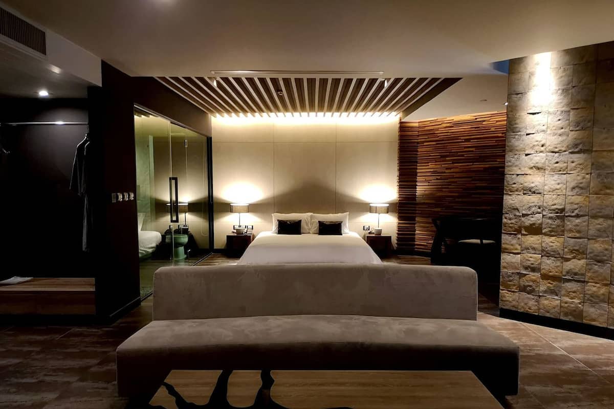 Quarto de hotel moderno com uma cama grande, mesas de cabeceira, um banco, uma parede de destaque em pedra e um espelho.