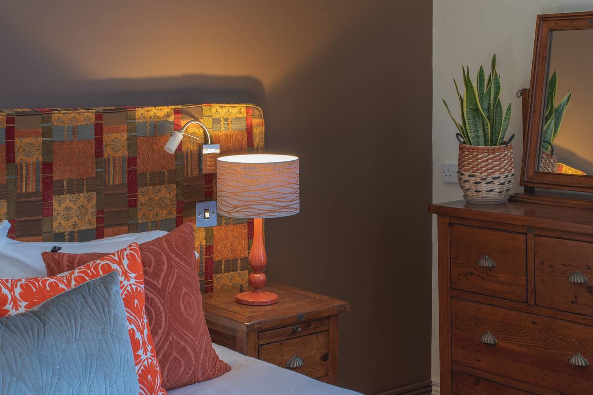 A neatly made bed with a pink quilt, a wooden nightstand with a lamp, and a dresser with a plant on top.