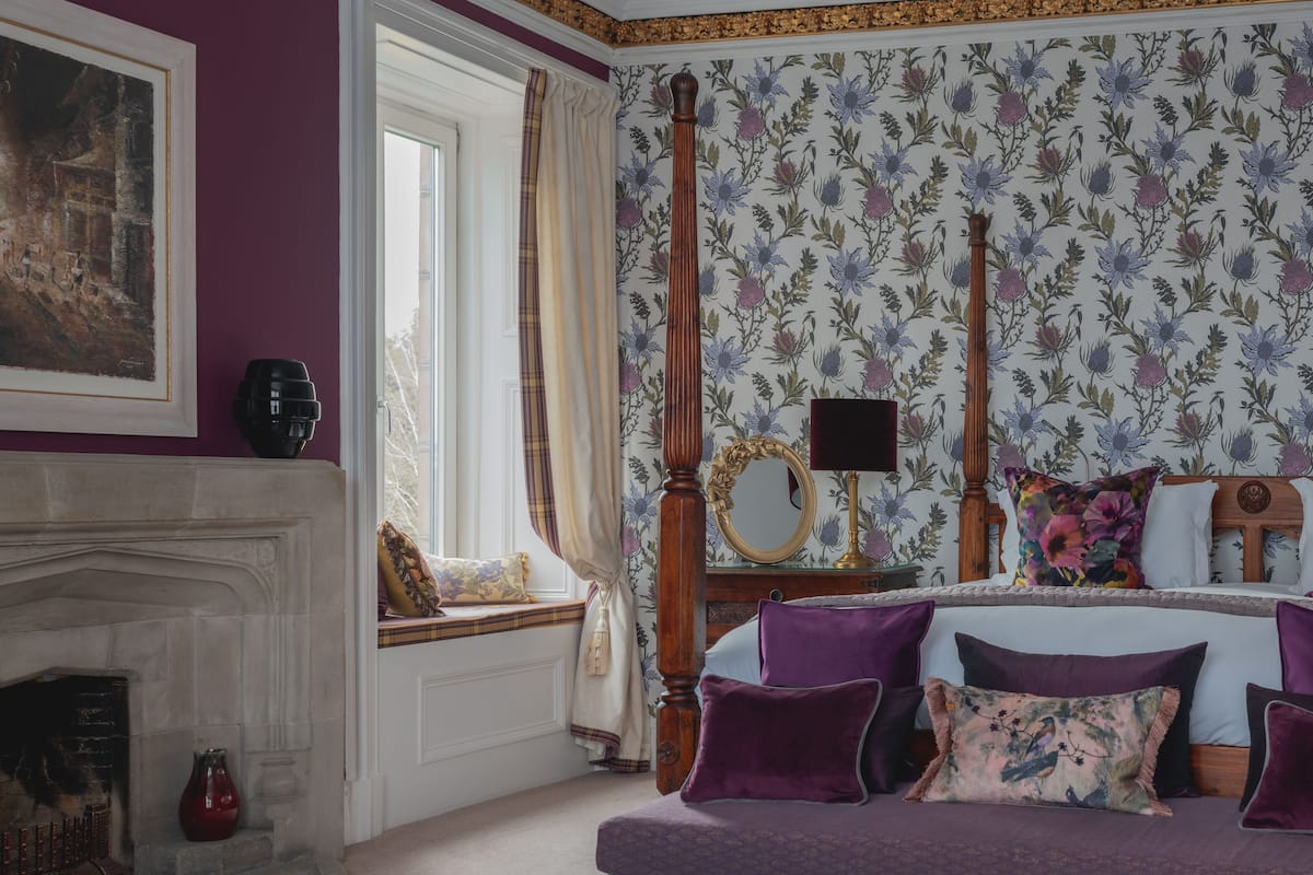 A bedroom with a fireplace, a bed with purple bedding, a floral wallpaper, and a window with curtains.
