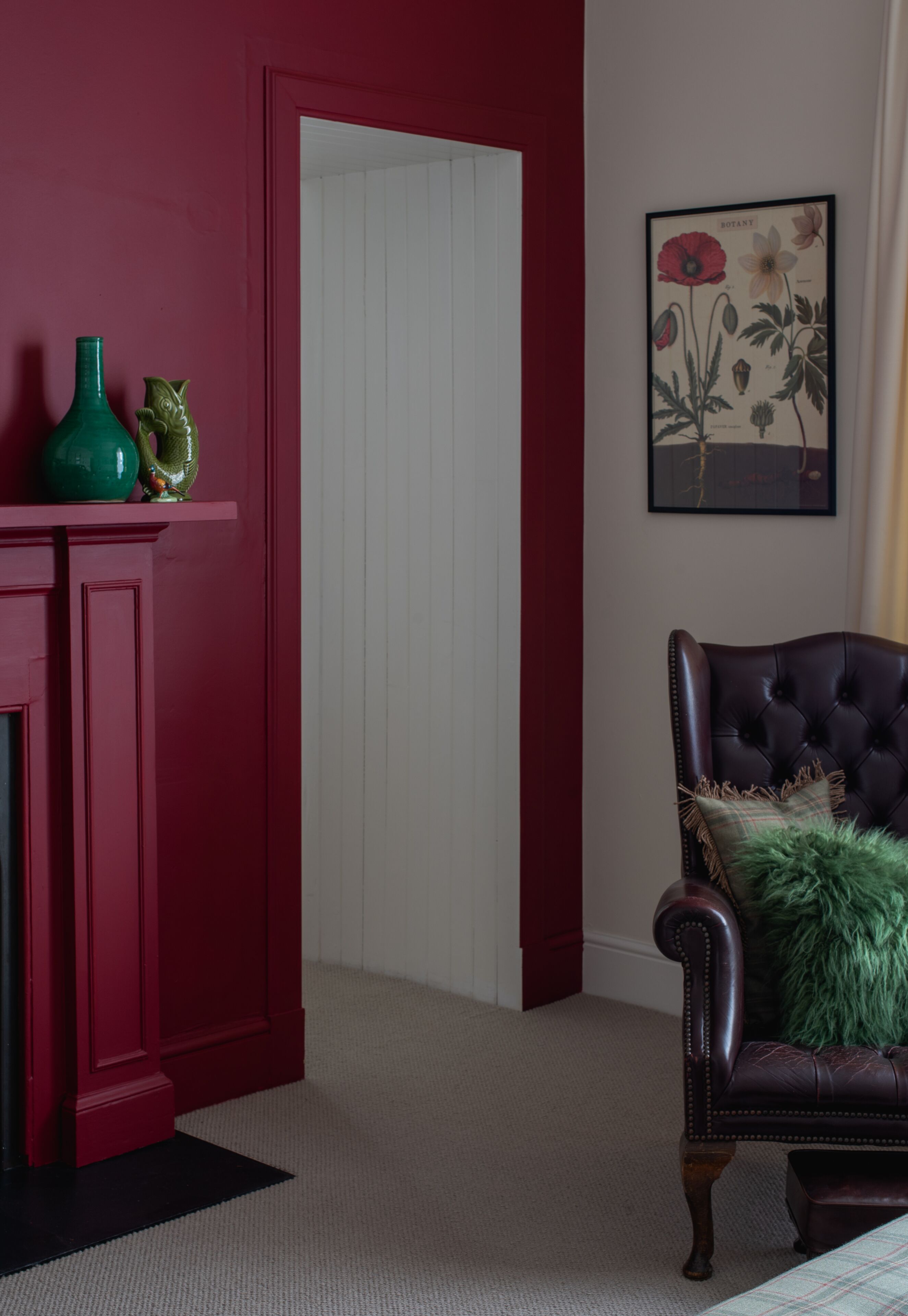 A room with a red door, a green vase, a framed botanical print, and a tufted leather chair with a green pillow.