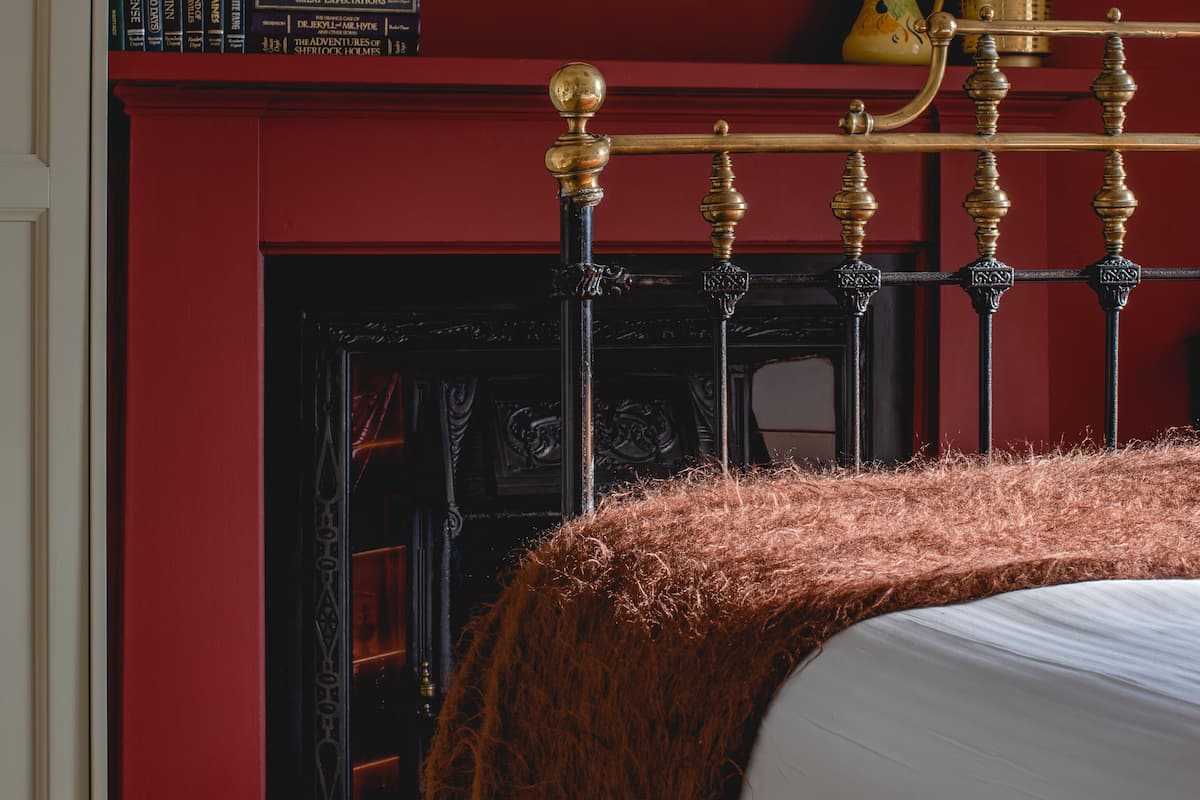 A bedroom with a red wall, a framed botanical print, a bookshelf, a yellow bowl, a brass bed frame, and a fireplace.