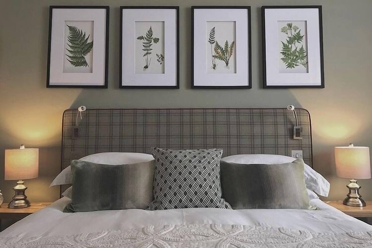 A neatly made bed with a plaid headboard, two bedside lamps, and four framed botanical prints on the wall.