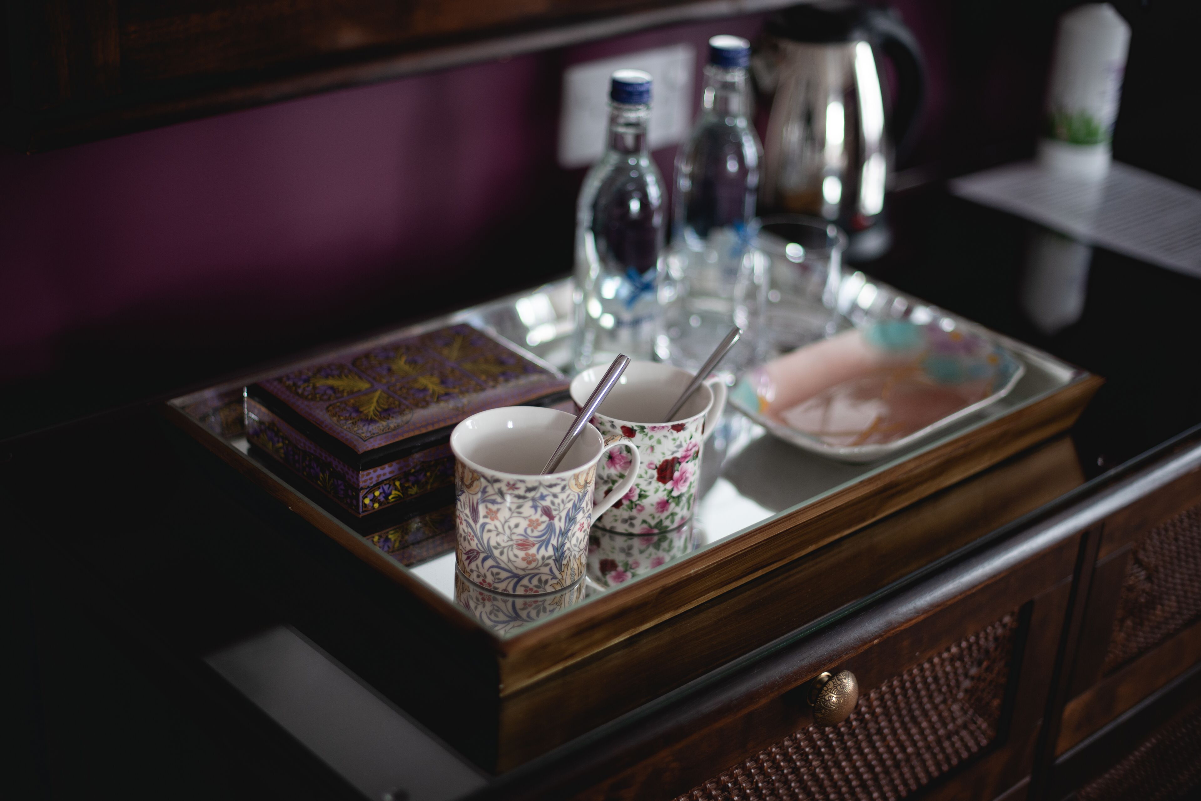 A tray with a box, two floral mugs, a plate with a spoon, and two bottles on a wooden surface.