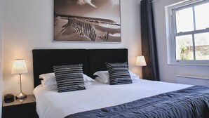 Een netjes opgemaakt bed met gestreepte kussens, een groot fotolijstje erboven en een raam met gordijnen.