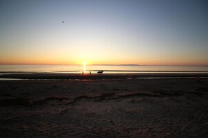 Praia ao pôr do sol, com céu claro, mar calmo e montanhas ao longe.