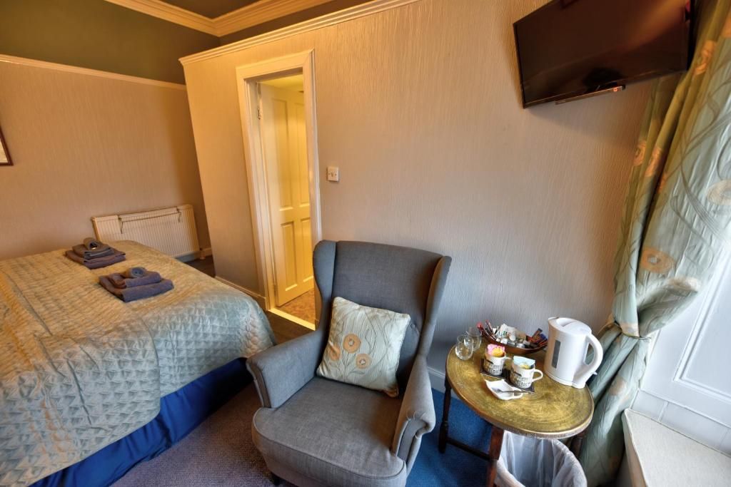 A hotel room with a bed, a chair, a small table with a kettle, and a TV mounted on the wall.