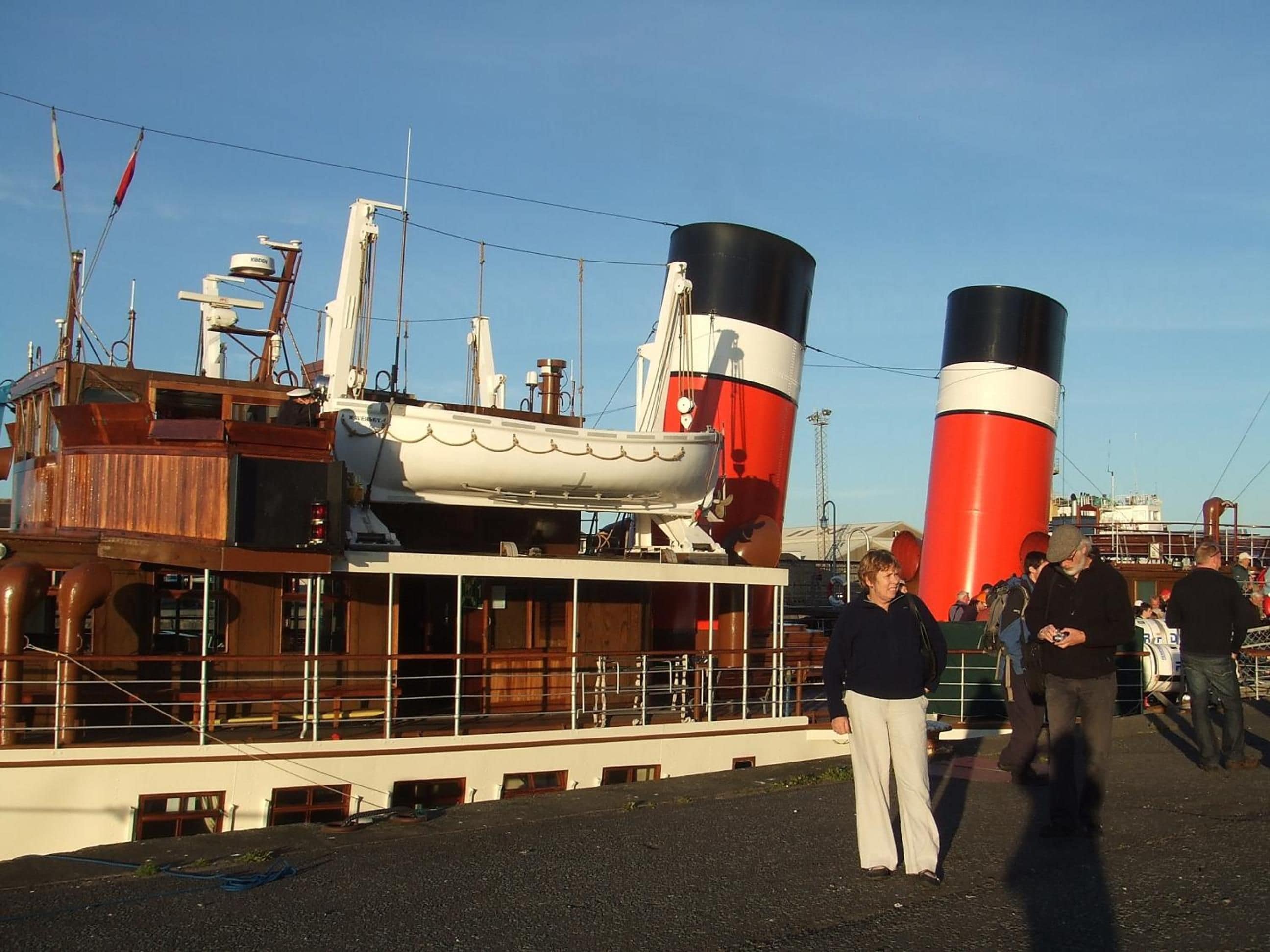 A large ship with two prominent smokestacks, a wooden deck, and a crowd of people.