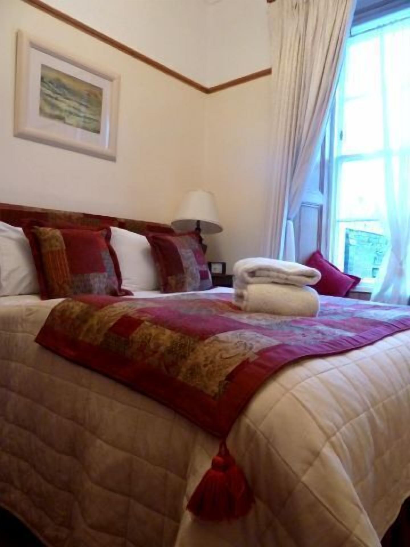 A neatly made bed with a red and beige quilt, a white pillow, and a red throw pillow. There is a white lamp on a bedside table and a framed picture on the wall.