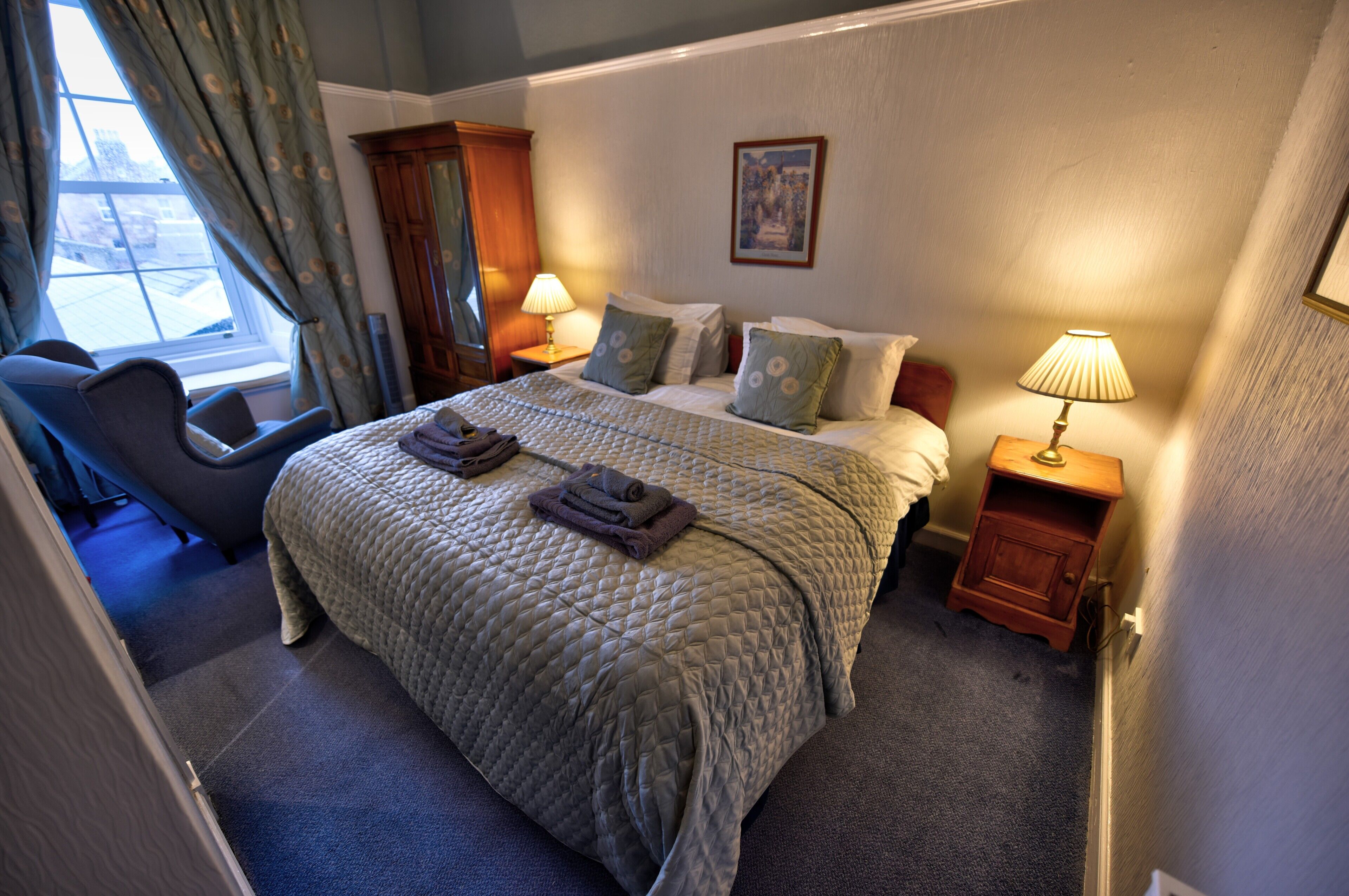 Une chambre d’hôtel avec un grand lit, deux tables de chevet avec des lampes, une chaise et une fenêtre avec des rideaux.
