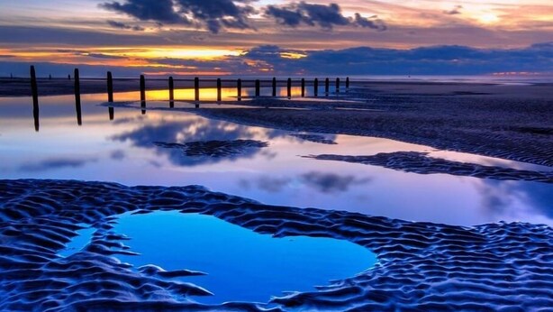 Ein Strand mit einem hölzernen Steg, der sich in ruhiges Wasser erstreckt, umgeben von Dünen.