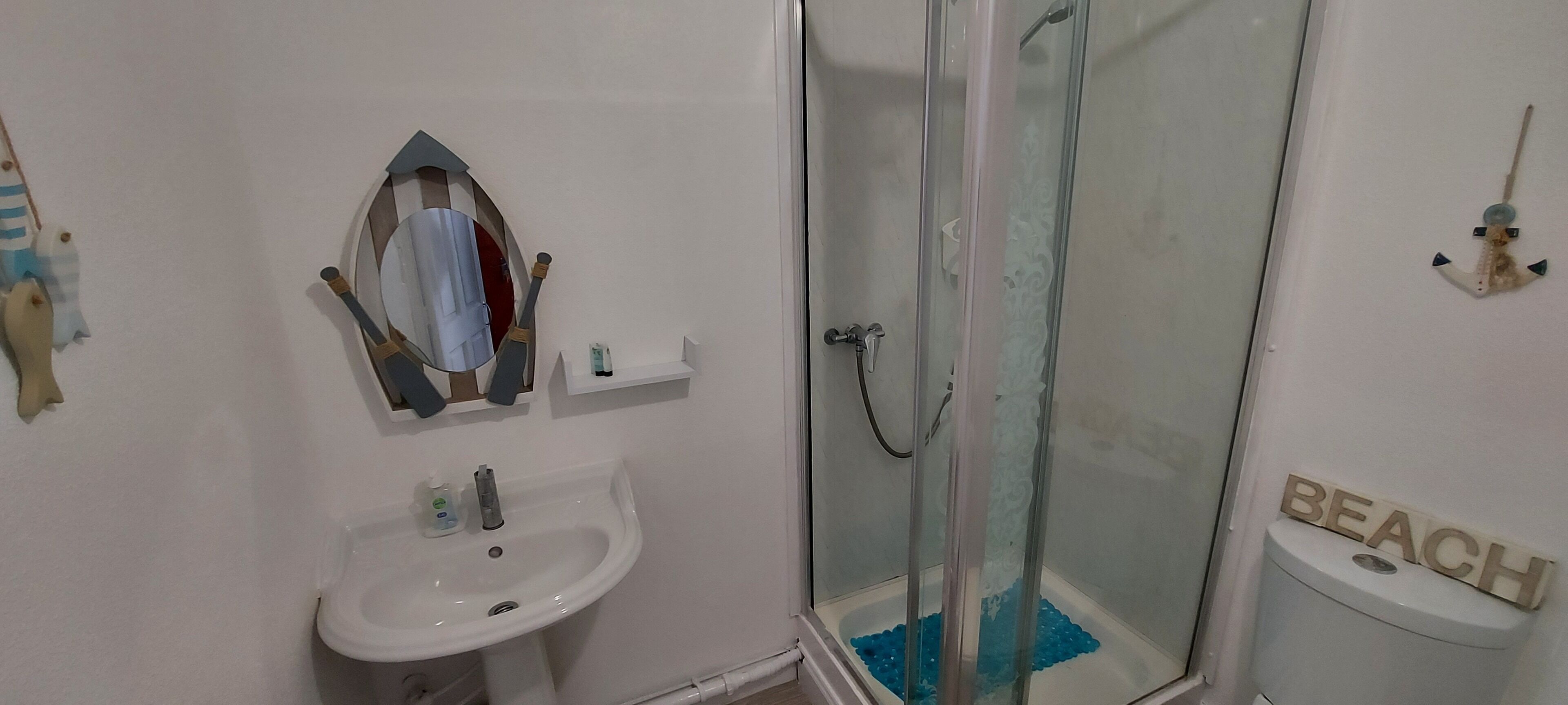 Un bany amb un lavabo blanc, un mirall, un box de dutxa de vidre i un vàter amb un rètol que diu 'BEACH'.