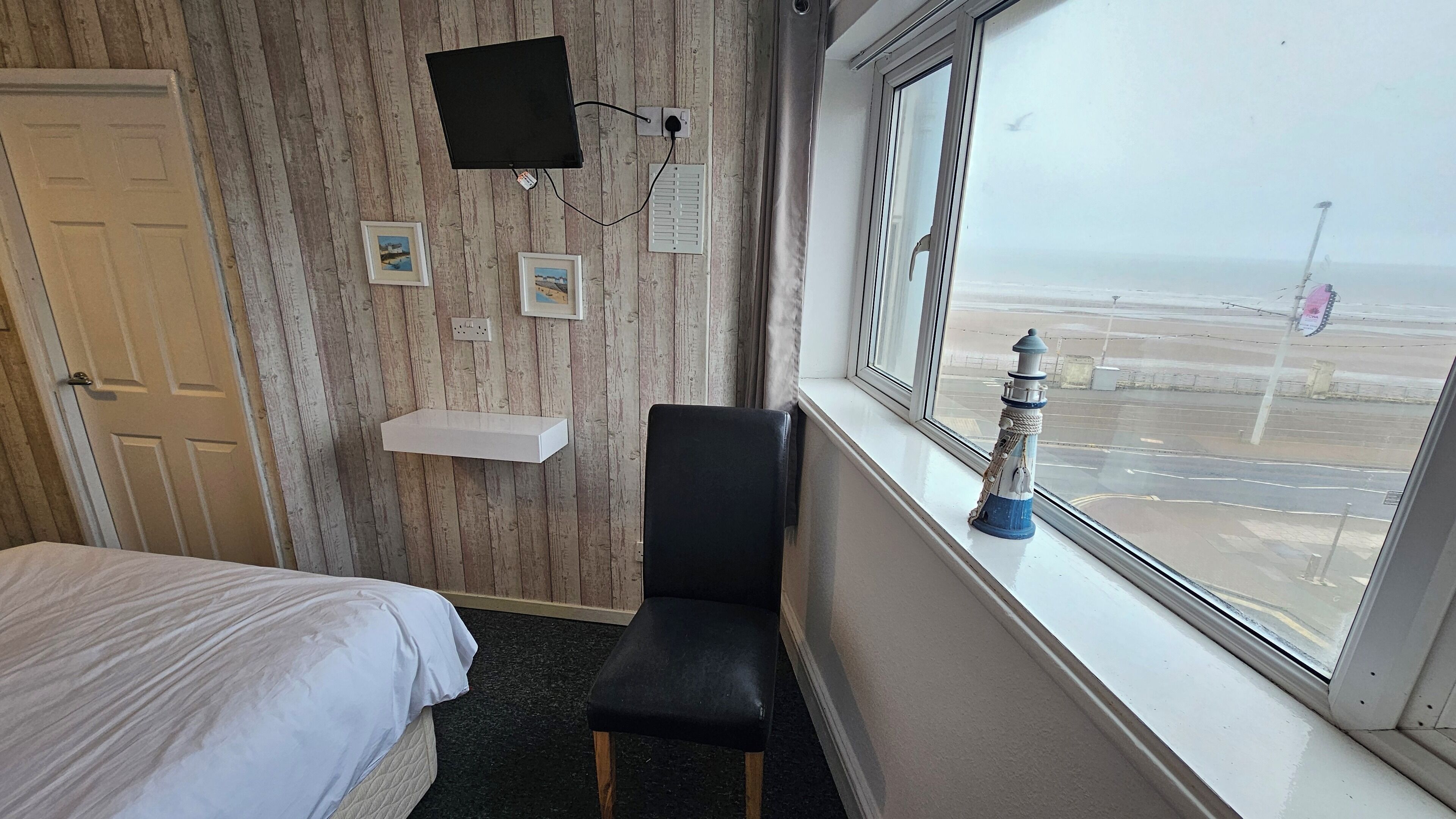 A hotel room with a bed, a black chair, a TV mounted on the wall, a small shelf, and a window with a view of the beach.