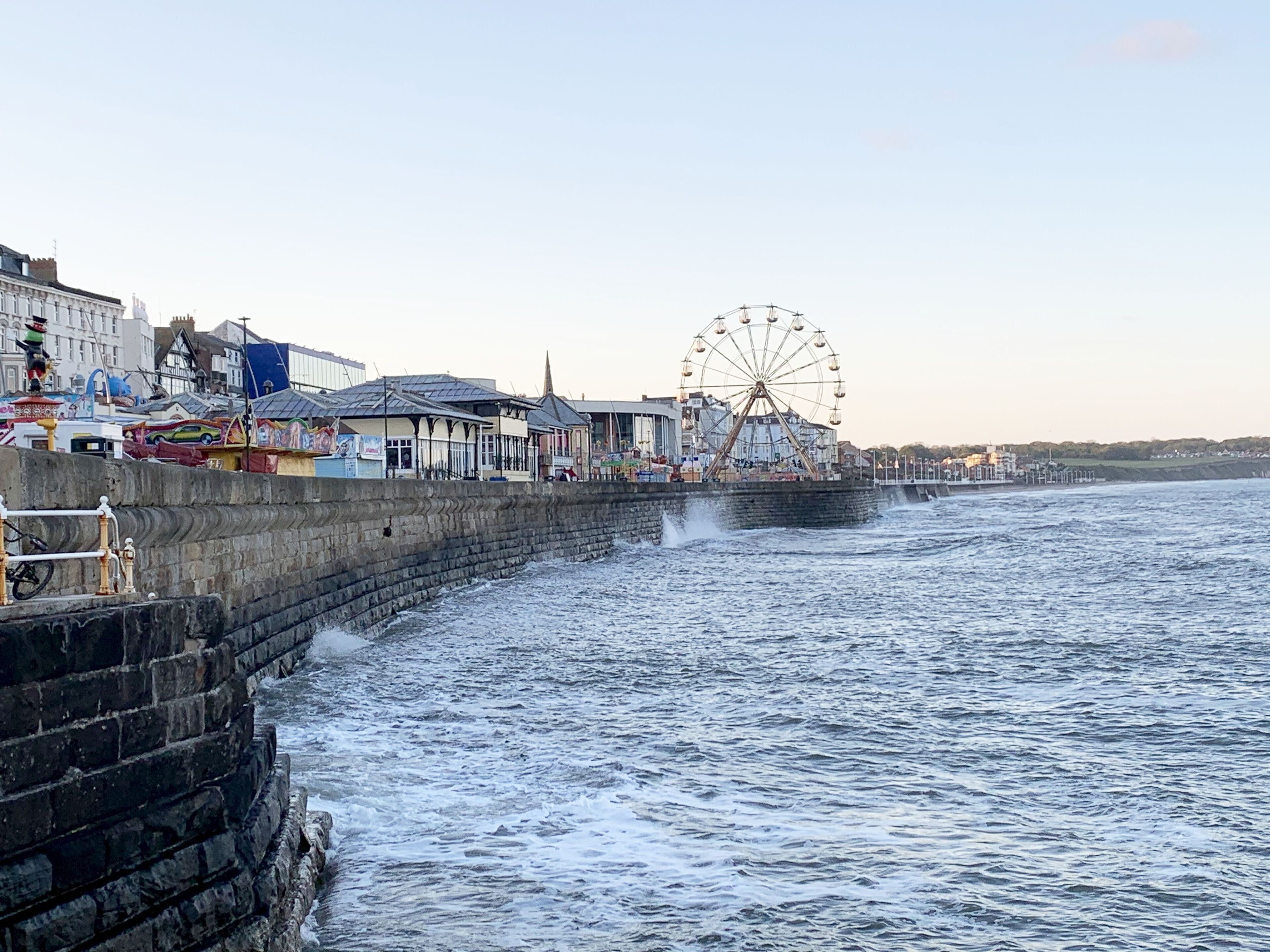 Une promenade en bord de mer agrémentée d’une grande roue, d’un quai en pierre et de bâtiments qui bordent la côte.