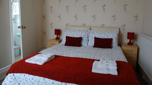 Ein ordentlich bezogenes Bett mit roter Decke und weißen Handtüchern, zwei rote Lampen auf Nachttischen und einem blumigen Wandpapier-Muster.