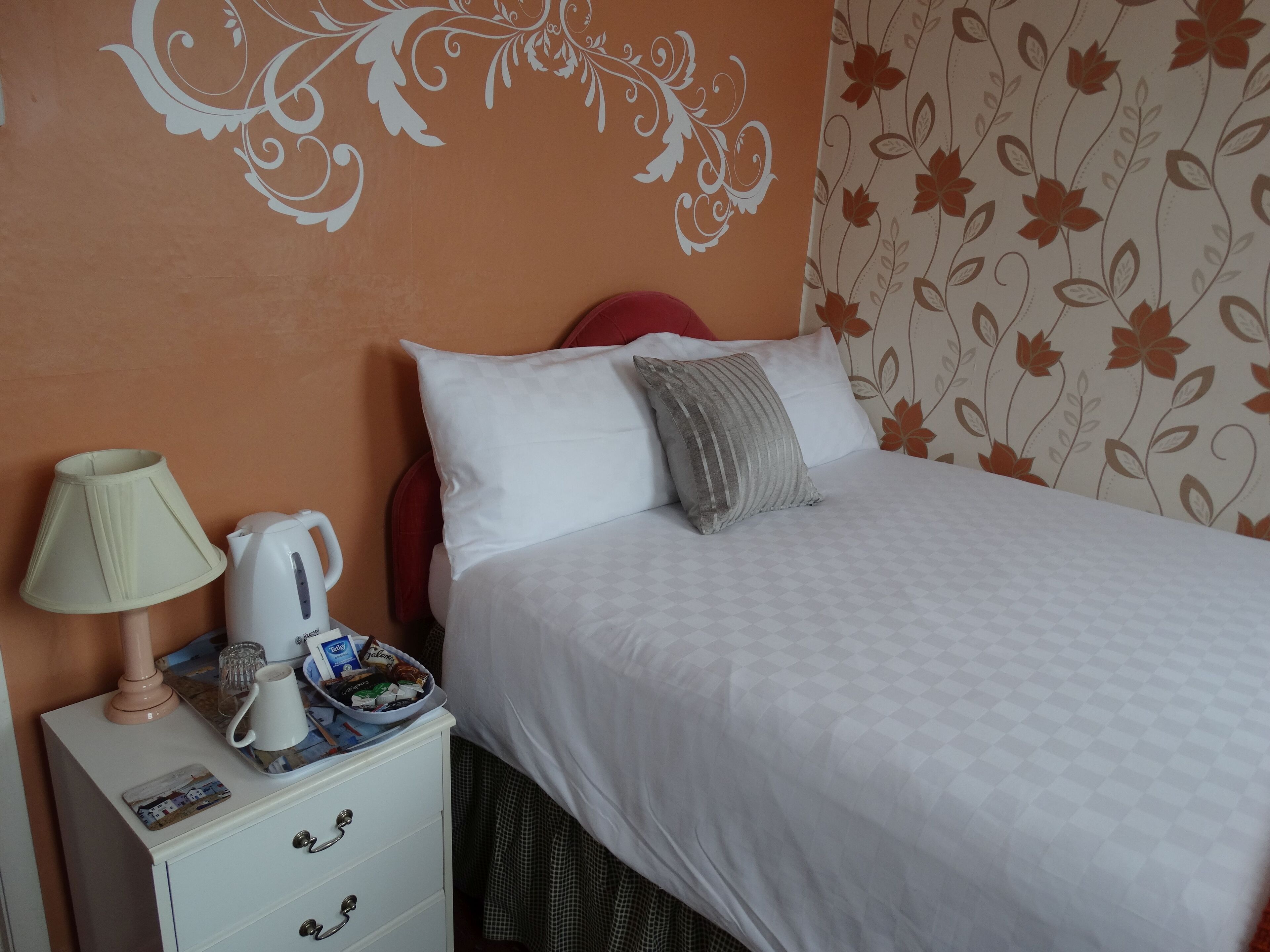 Una cama bien hecha con ropa de cama blanca y una almohada gris, una mesita de noche blanca con una lámpara, un hervidor y una bandeja de aperitivos.