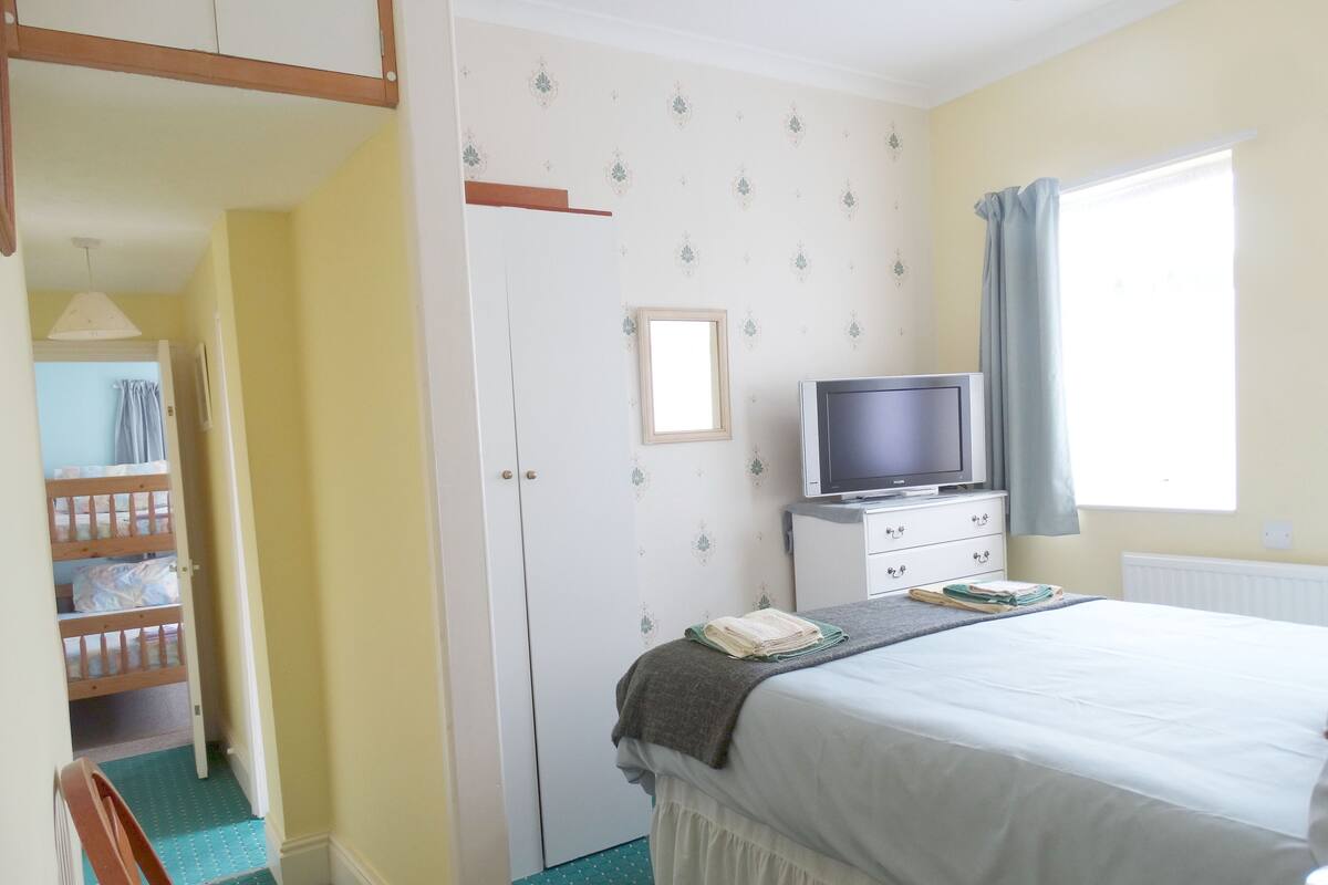 A bedroom with a bed, a television on a dresser, a window with curtains, and a door leading to a bathroom.