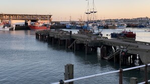 Ein Hafen mit mehreren Booten, die an der Kaimauer festgemacht sind, einer Brücke im Hintergrund und einem Gebäude mit einem Balkon mit Blick auf das Wasser.