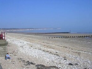 A beach with a clear sky, a long pier extending into the water, and a person sitting on the sand.