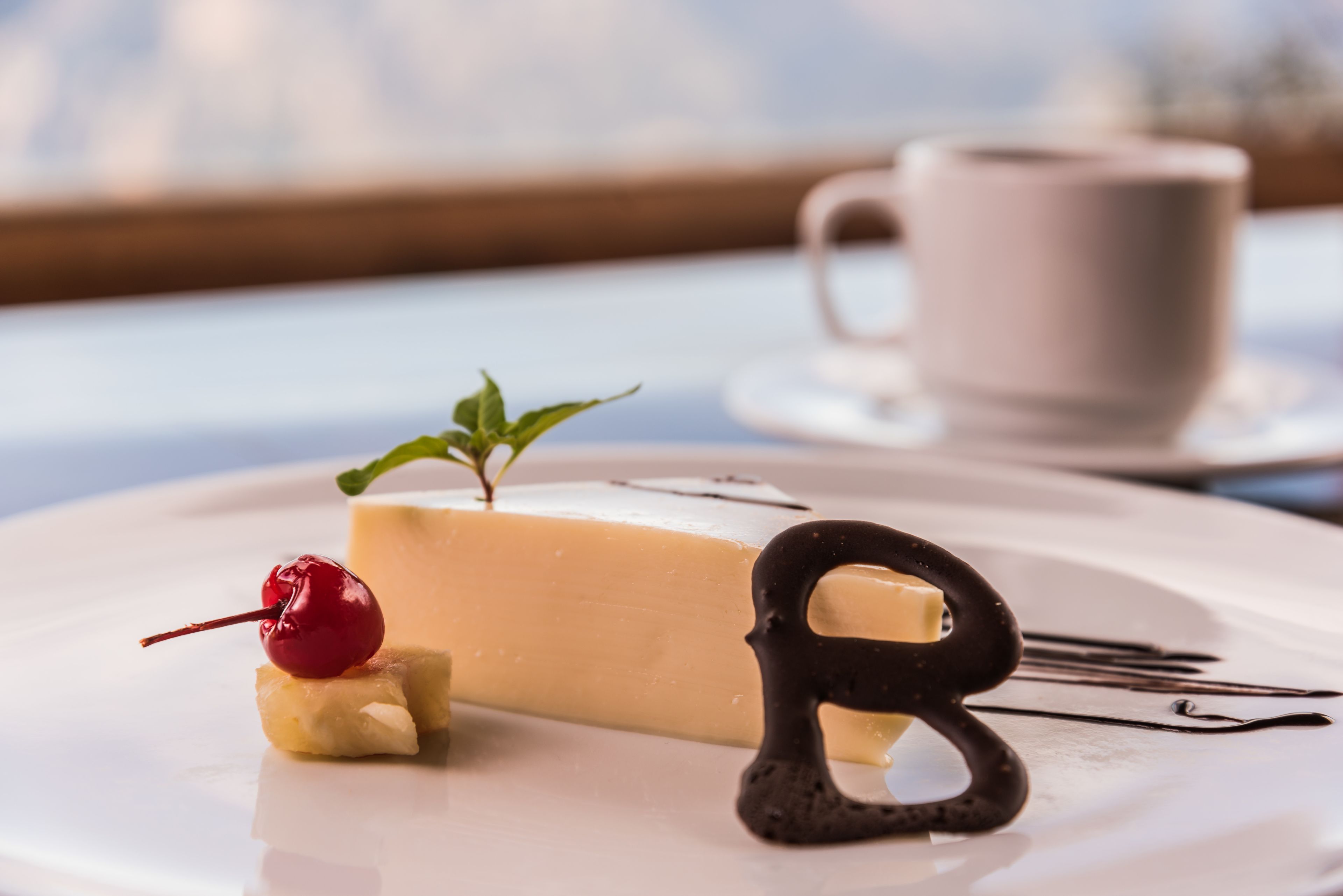 Dessert op een bord met een kers en munt, een chocolade letter 'B' en een kopje op de achtergrond.
