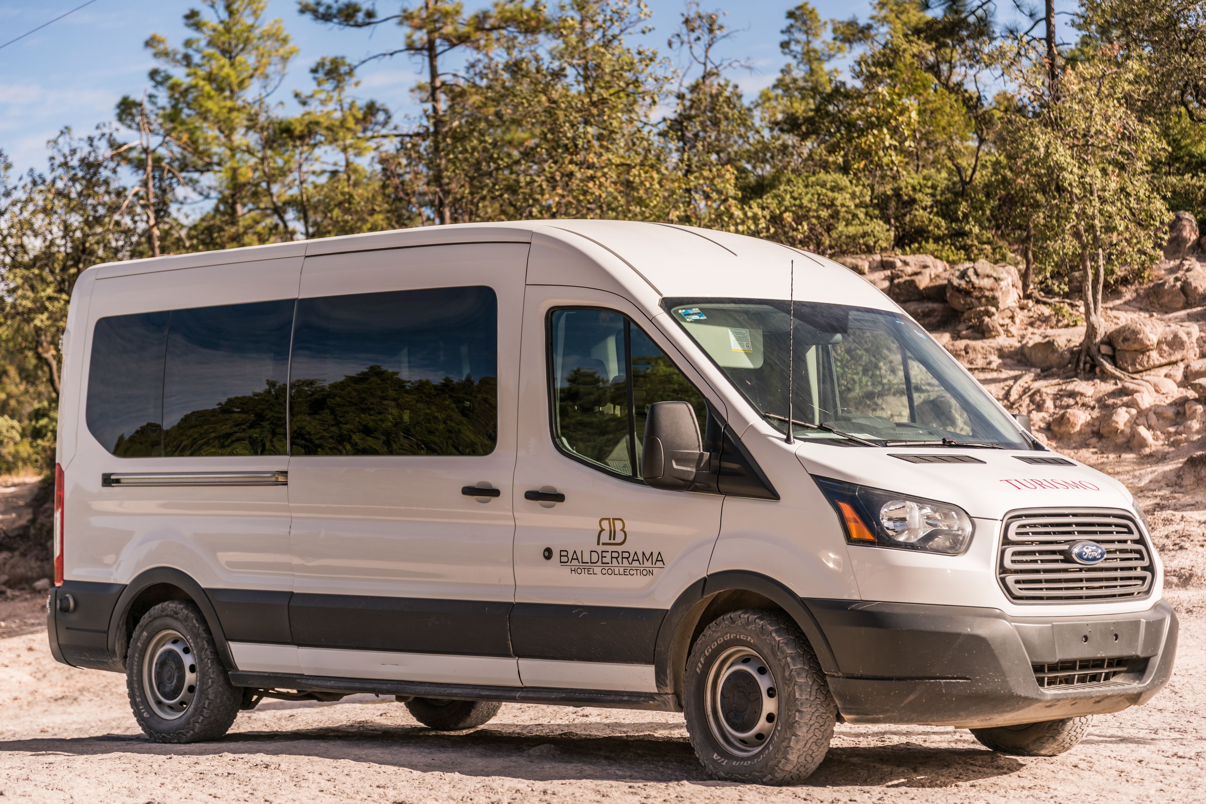 Una camioneta blanca con el logotipo de 'Balderrama Hotel Collection', estacionada en un camino de tierra con terreno rocoso y árboles de fondo.
