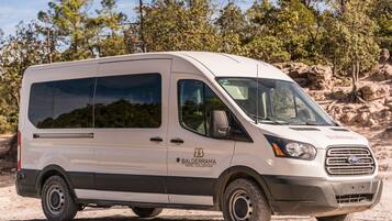 Una camioneta blanca con el logotipo de 'Balderrama Hotel Collection', estacionada en un camino de tierra con terreno rocoso y árboles de fondo.
