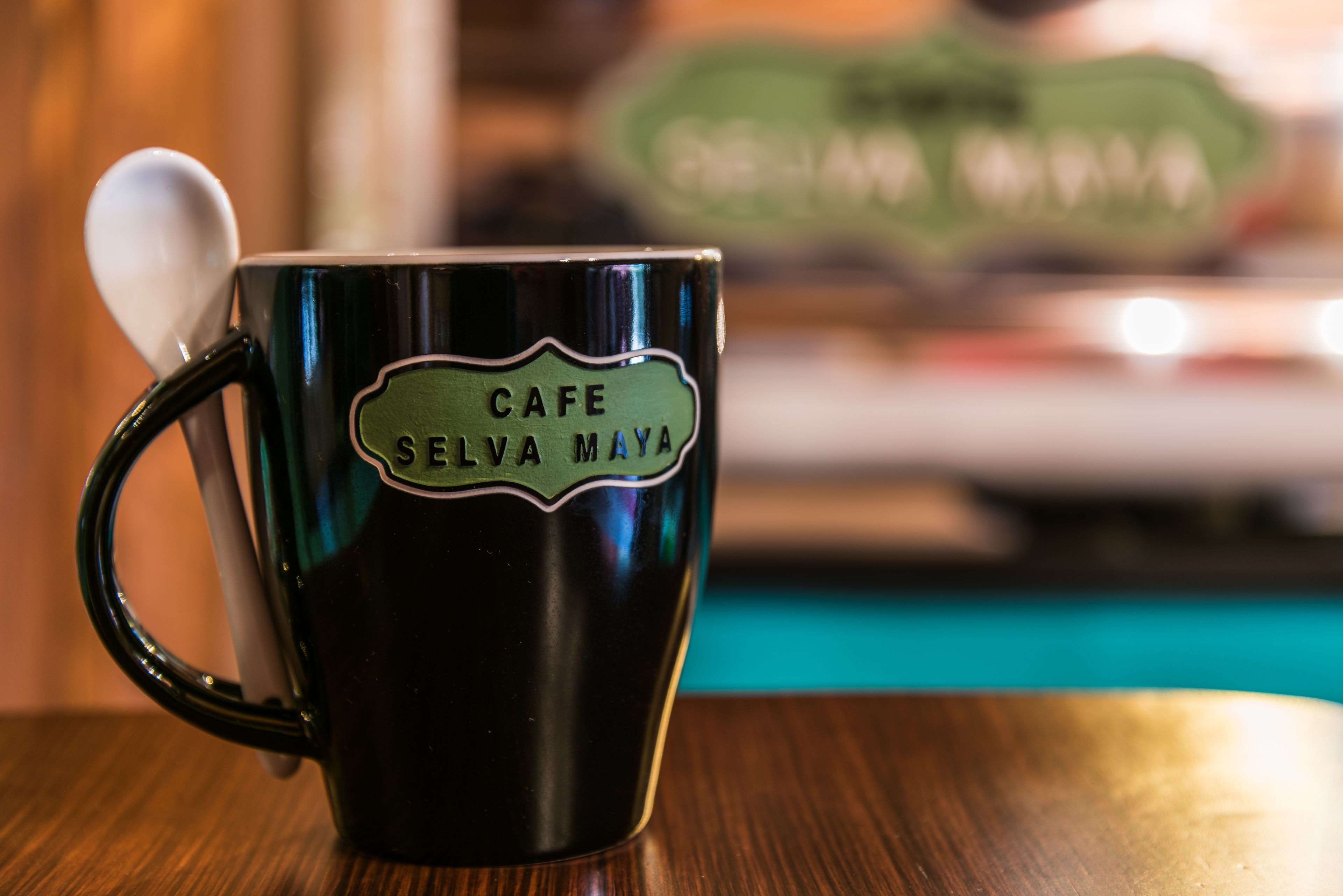 Una taza verde oscuro con una cuchara, con la etiqueta 'CAFE SELVA MAYA', sobre una mesa de madera.