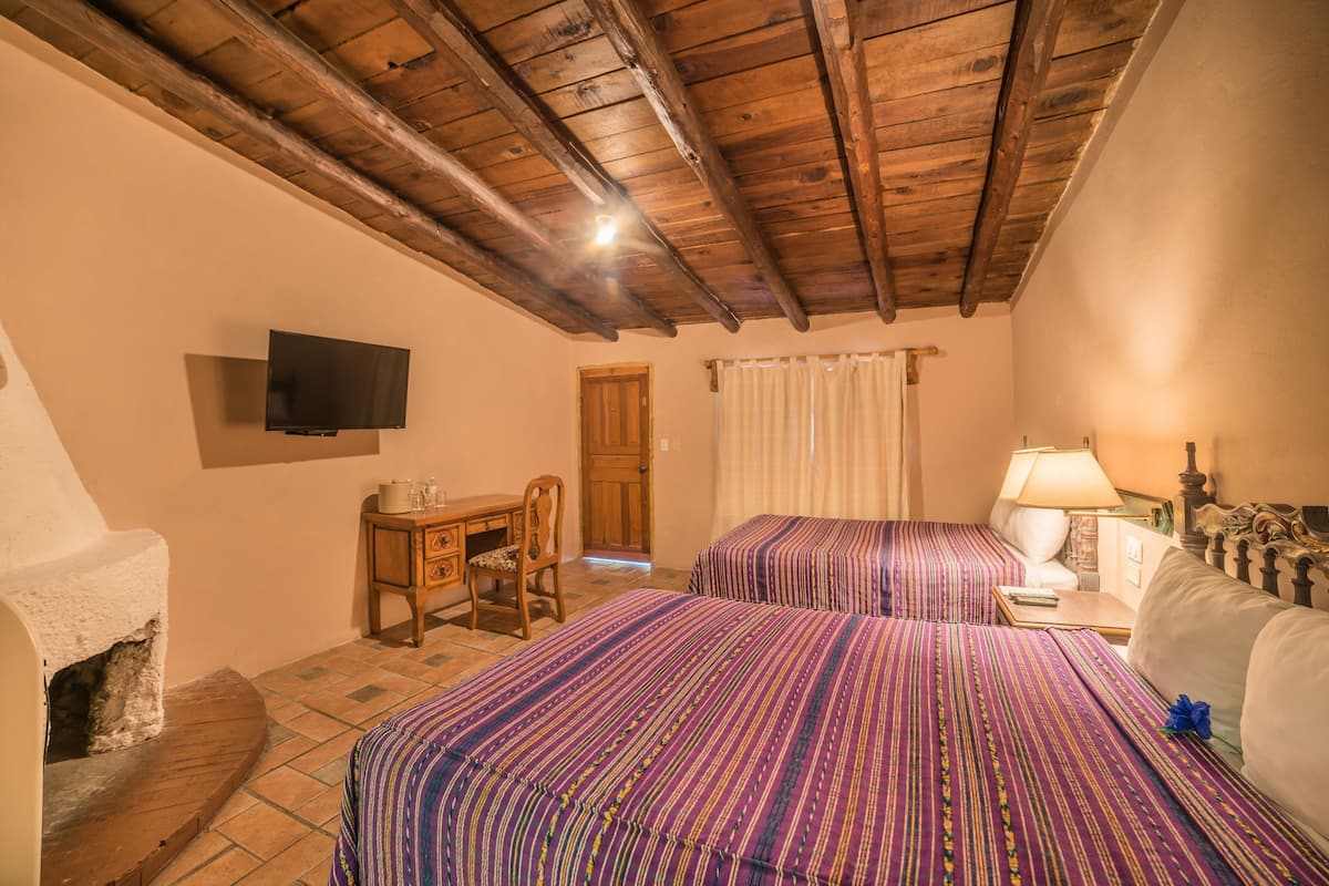 Een kamer met houten plafond, een aan de muur bevestigde televisie, twee bedden met streepjesbeddengoed, een bureau met stoel en een open haard.