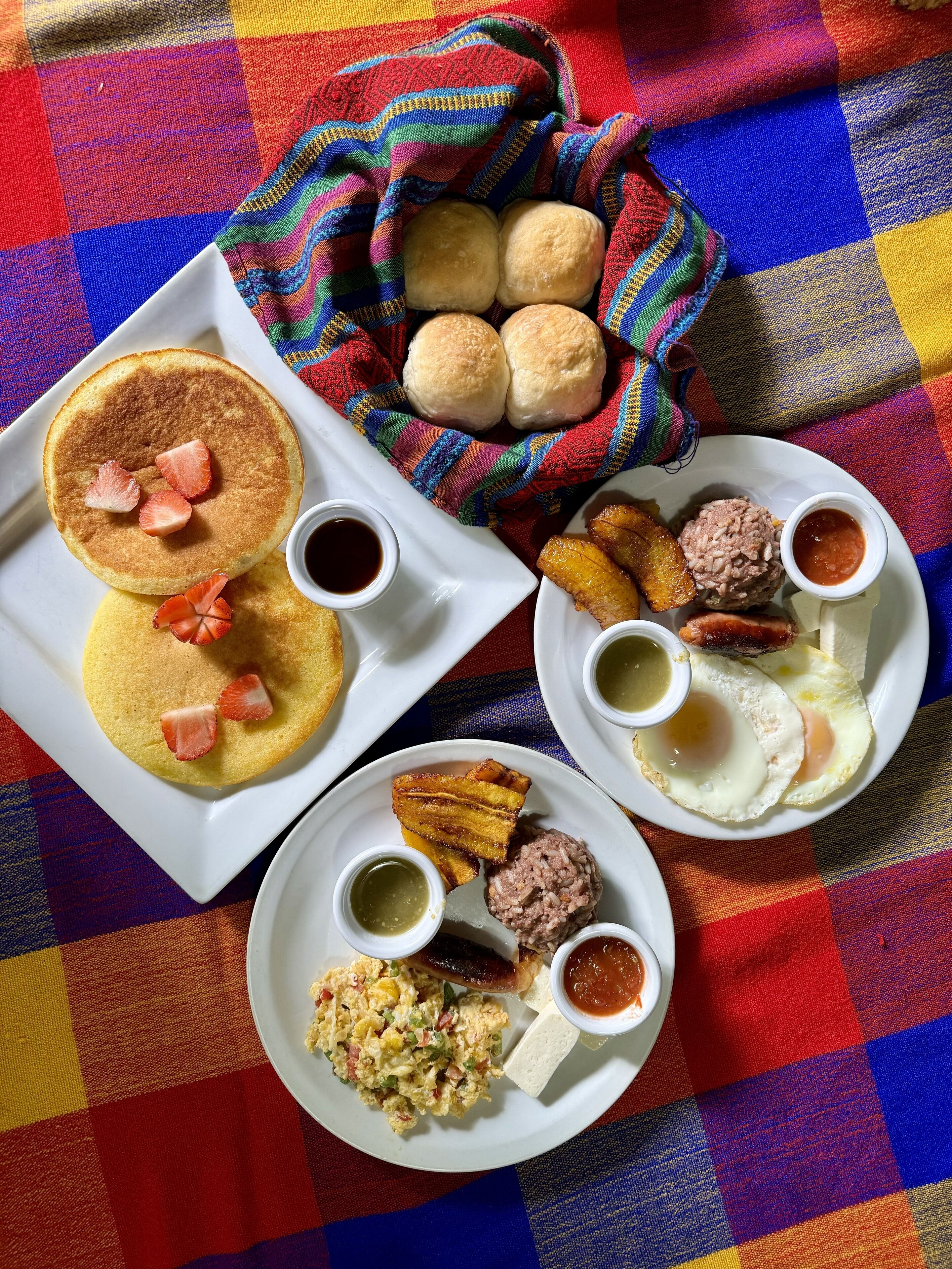 Un desayuno abundante con tortitas, huevos, salchichas y diferentes acompañamientos sobre platos blancos, sobre un mantel a cuadros coloridos.