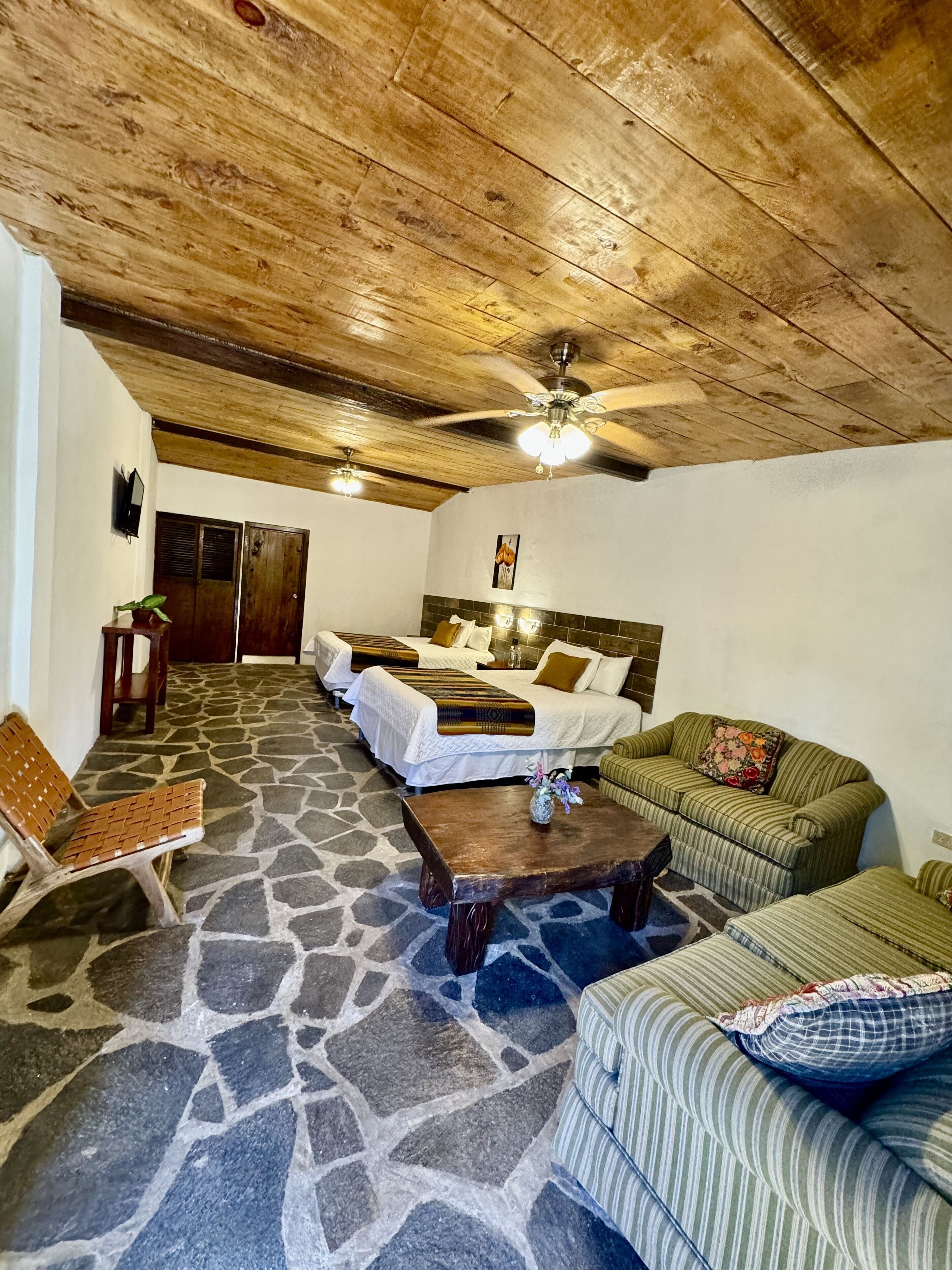 A room with a stone floor, wooden ceiling, two beds with white bedding, a green striped sofa, a wooden coffee table, and a television on the wall.