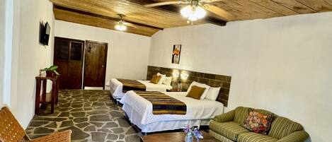 A room with a stone floor, wooden ceiling, two beds with white bedding, a green striped sofa, a wooden coffee table, and a television on the wall.