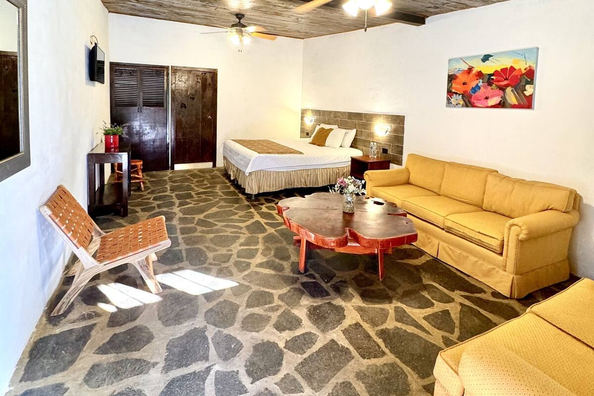 Una habitación con suelo de piedra, techo de madera, cama con edredón blanco, sofá amarillo, banco de madera y una mesa con un jarrón.