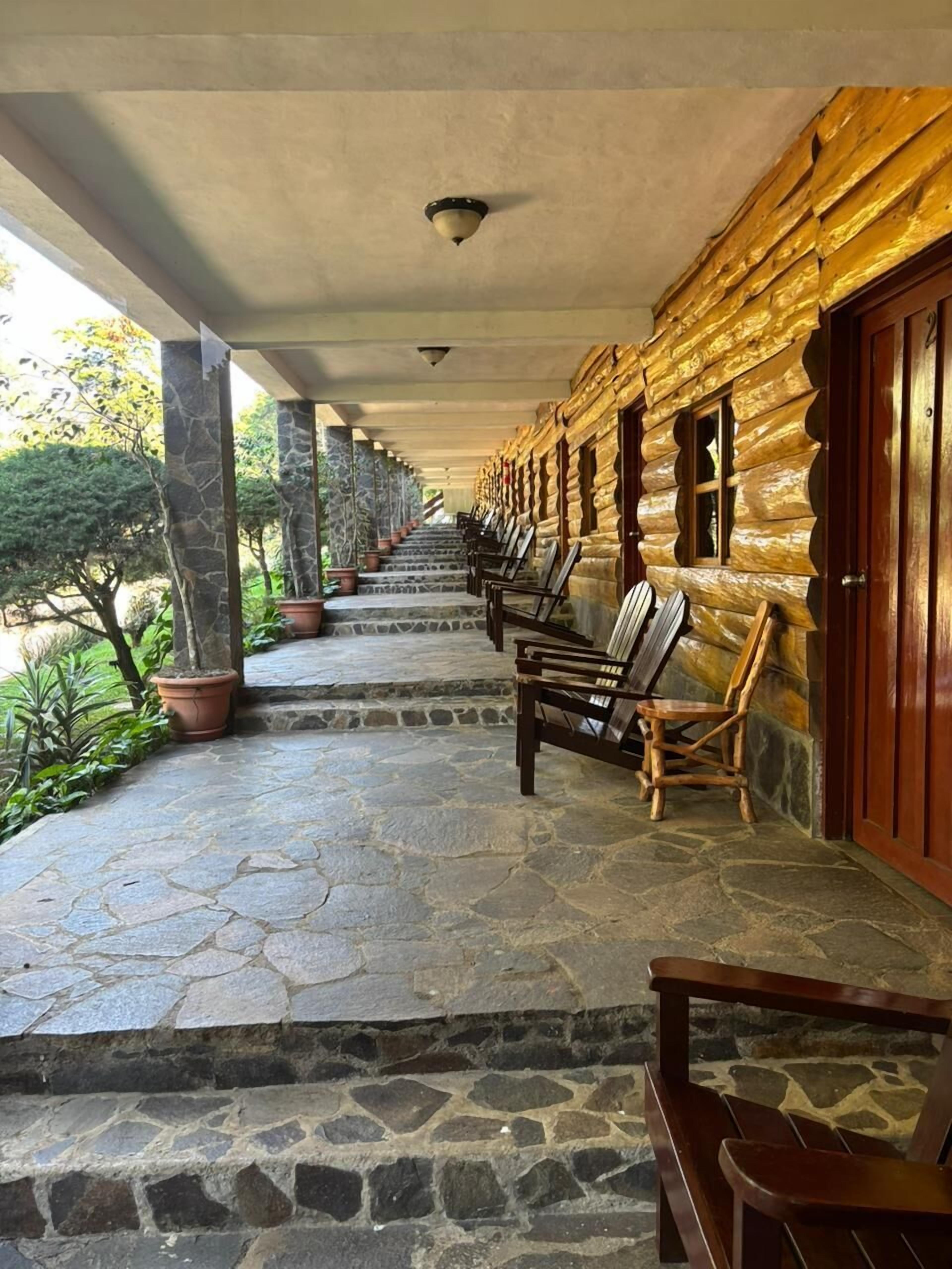 A covered outdoor area with stone flooring, wooden benches, and a row of potted plants.