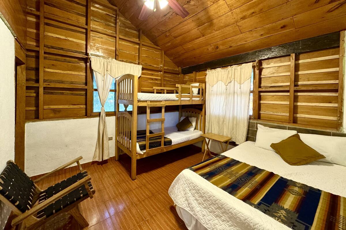 Habitación tipo cabaña con paredes de madera, una litera, una cama individual, ventilador de techo y ventana con cortinas.