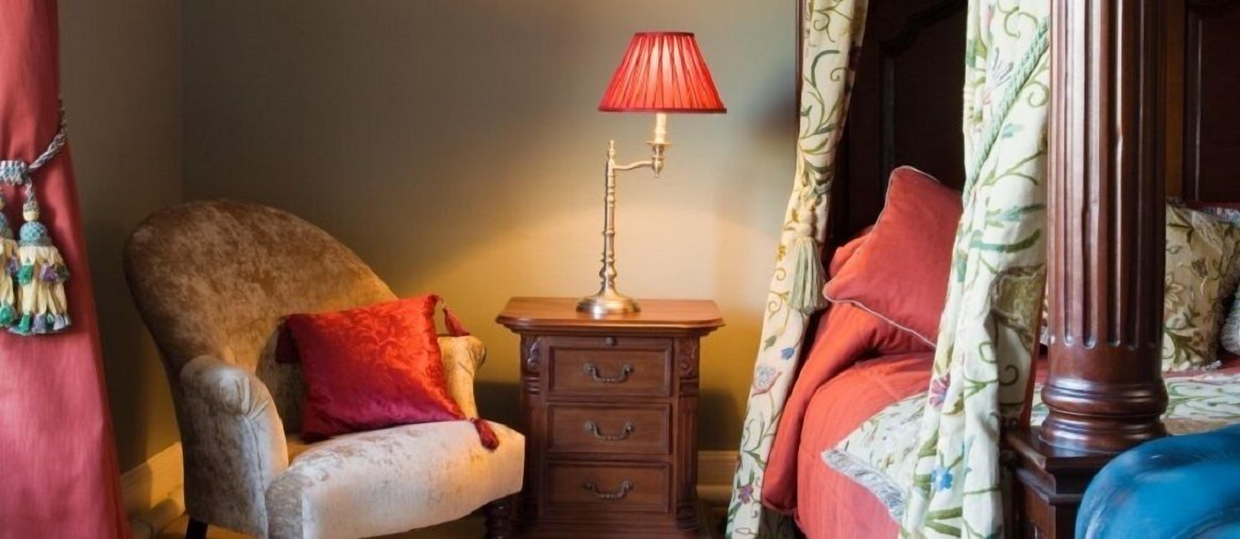 A bedroom with a wooden nightstand, a red lamp, a chair with a red pillow, and a bed with floral curtains.