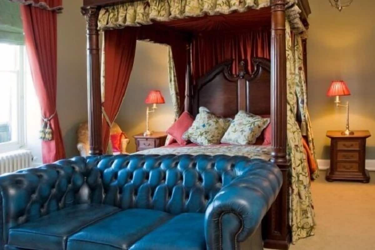 Ein Himmelsbett mit rotem und goldenem Baldachin, ein blaues Ledersofa und Blick auf Grünfläche draußen.