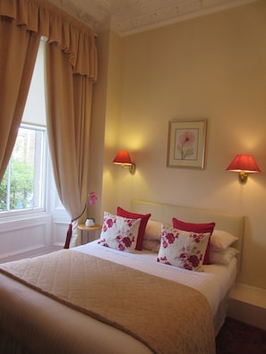 Una cama bien hecha con cubrecolcha acolchado, dos almohadas rojas y un cuadro con flores en la pared.