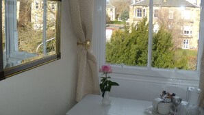 A bathroom with a white vanity, a window with a view of houses and trees, a mirror with a gold frame, and a vase with a single pink flower. - Sonas Guesthouse (Edinburgh)