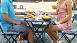 Dos personas sentadas en una mesa al aire libre, con una vista de un hotel y una cafetería al fondo.