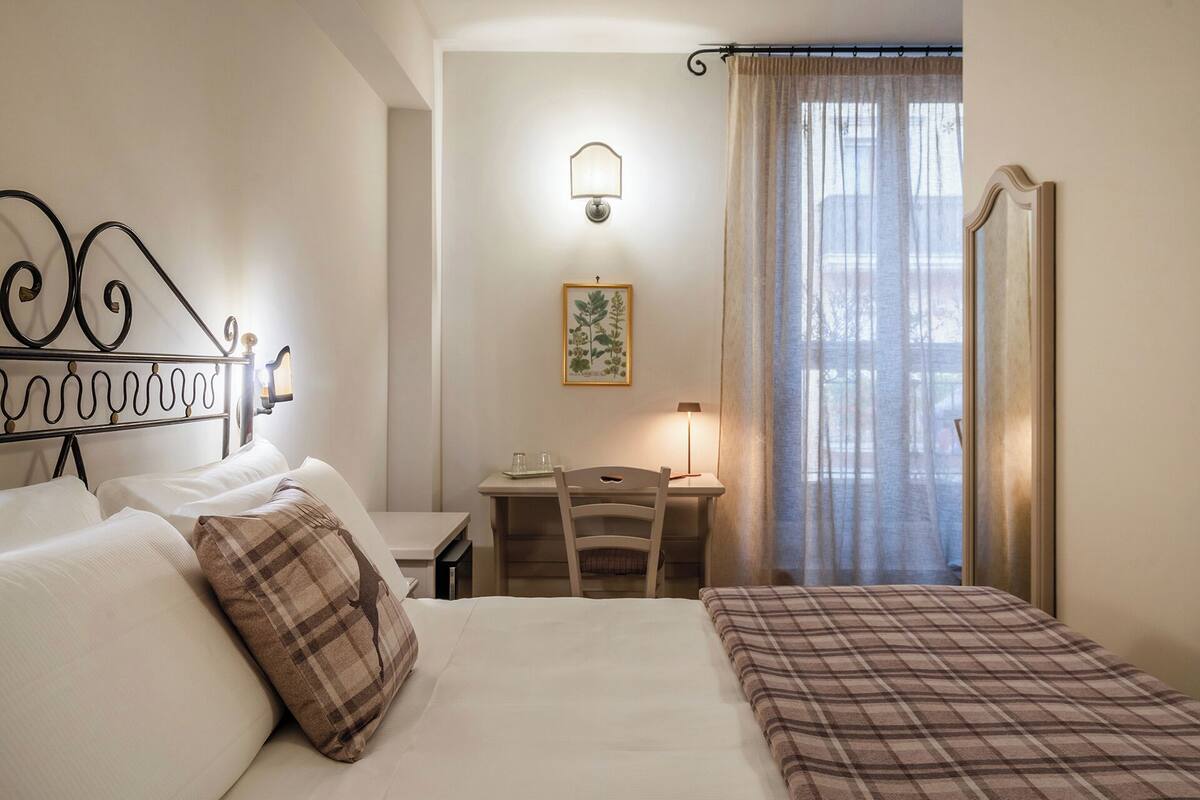 A hotel room with a bed, a desk, a chair, a lamp, and a window with curtains.