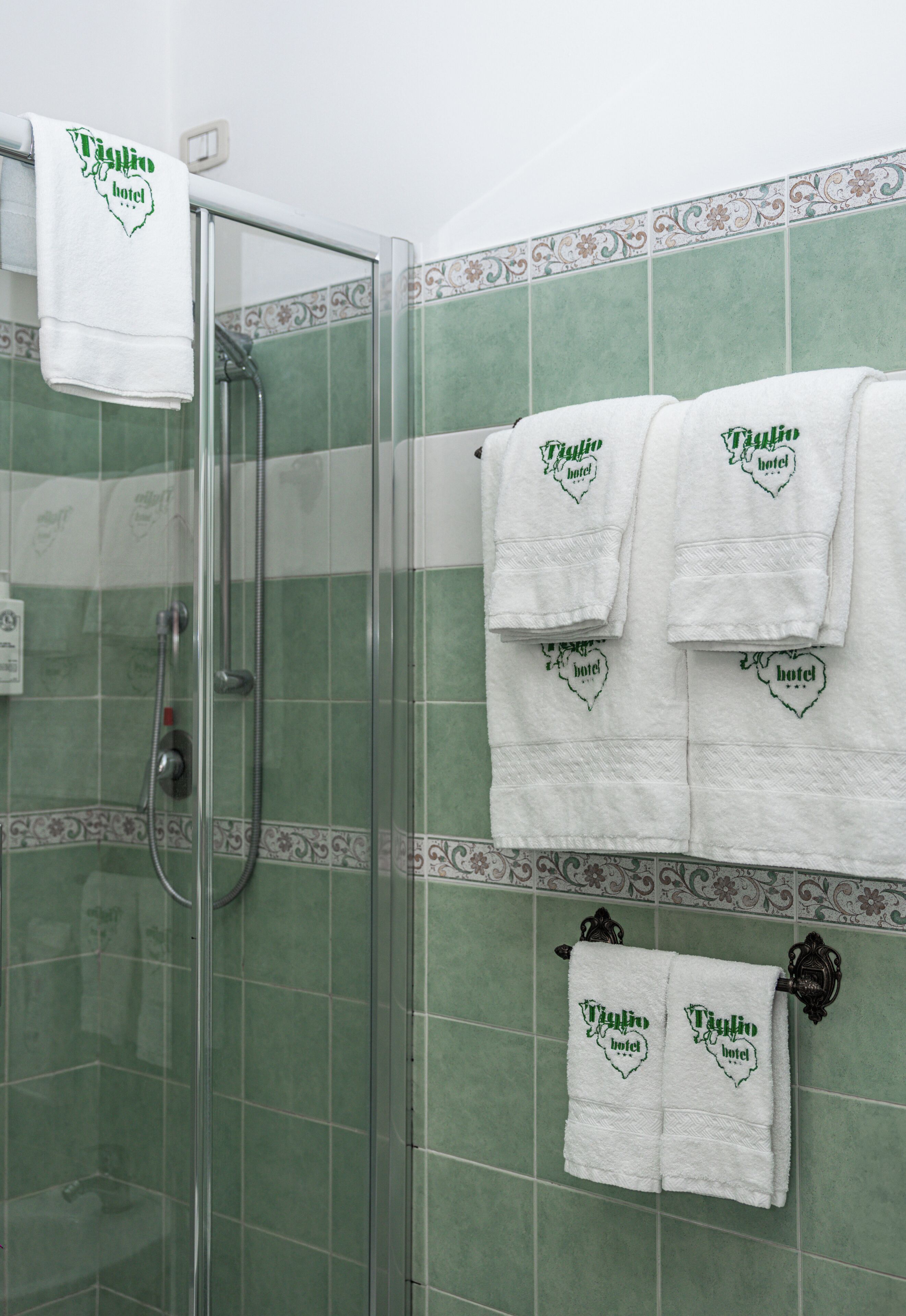 Un baño con azulejos verdes, cabina de ducha de vidrio y toallas blancas con la marca 'Villa Rio Hotel'.