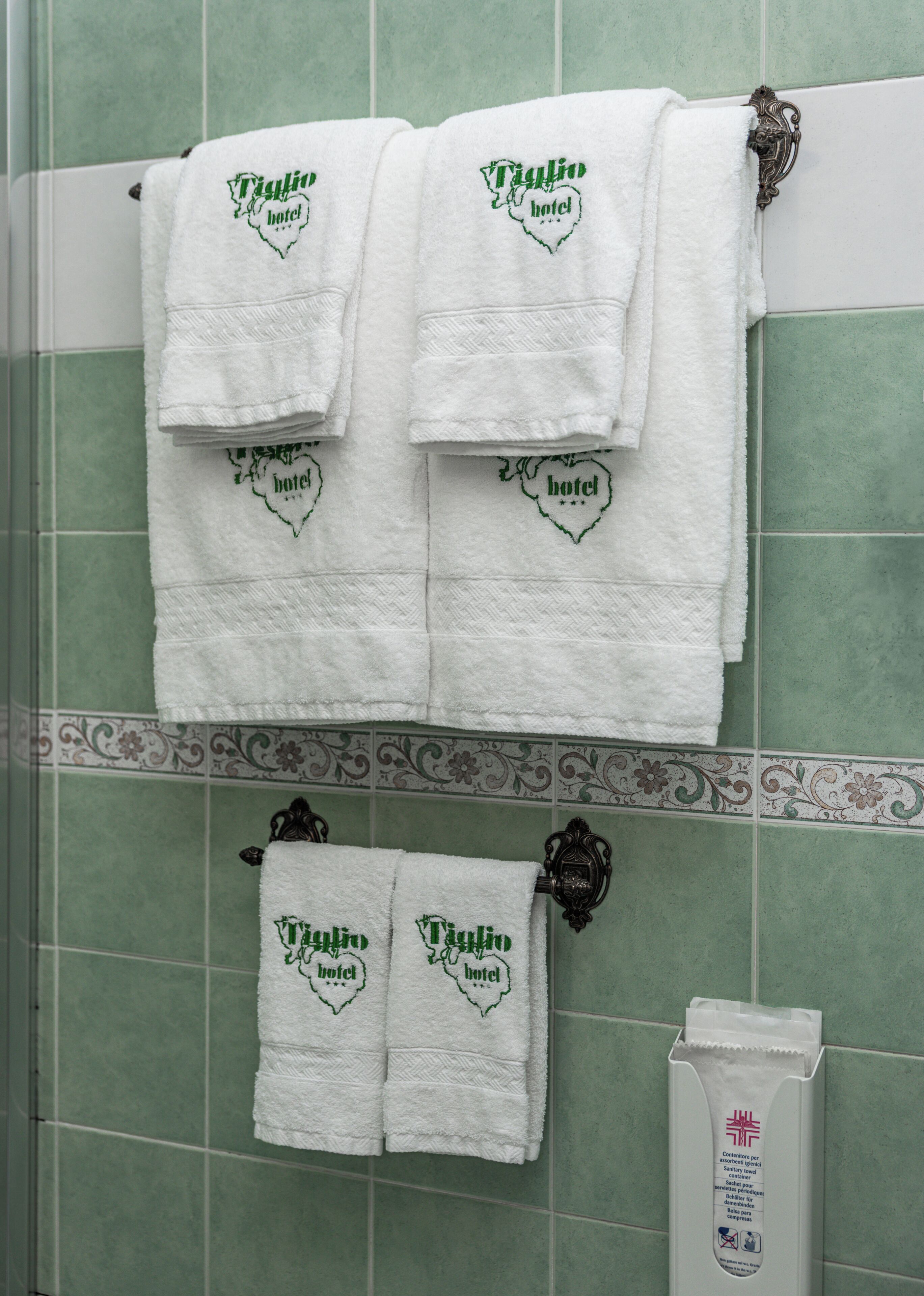 Un baño con azulejos verdes, dos toallas blancas con el bordado 'Taglio hotel' y un secador de manos.