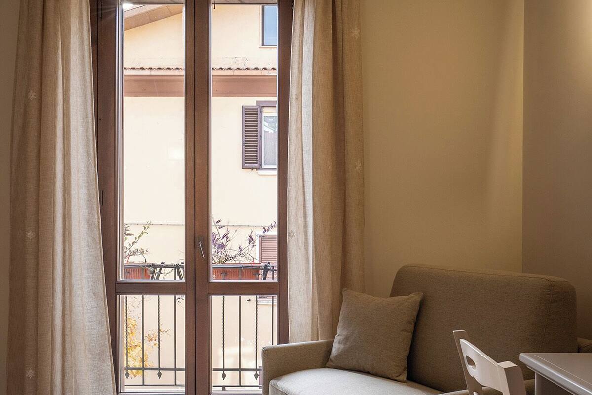 A small, cozy room with a sofa, a chair, and a bed. There is a window with curtains, and a balcony is visible outside.