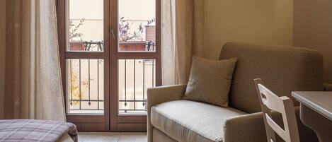 A small, cozy room with a sofa, a chair, and a bed. There is a window with curtains, and a balcony is visible outside.