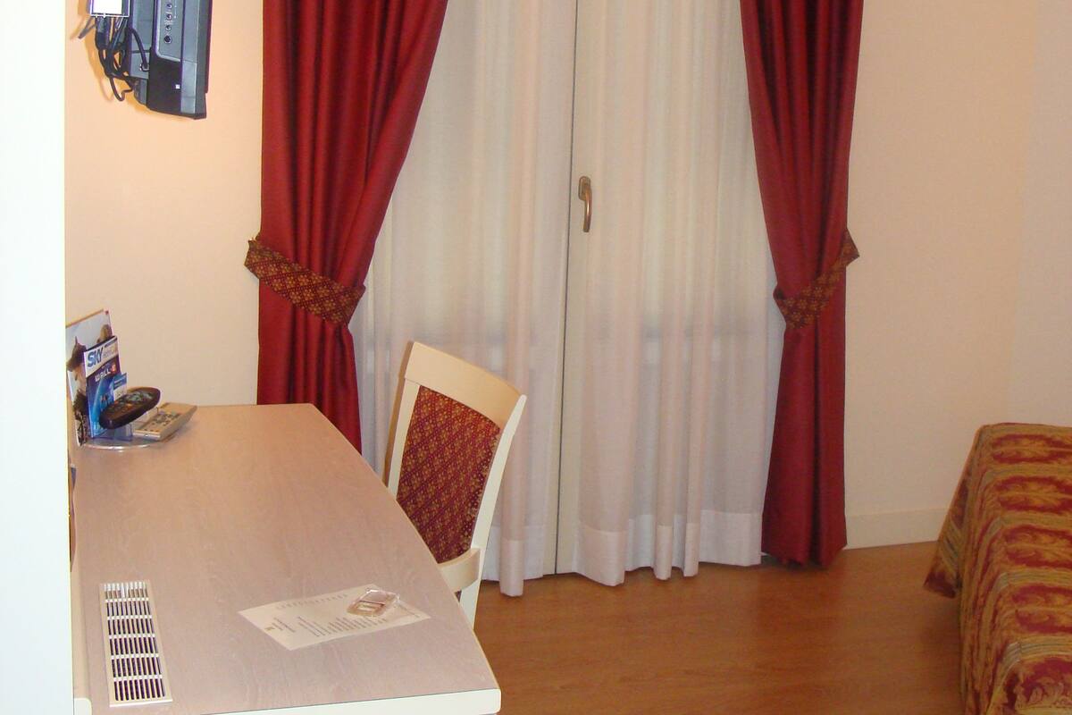 Habitació d'hotel amb un llit, una taula de treball amb una cadira, una televisió i una finestra amb cortines vermelles.