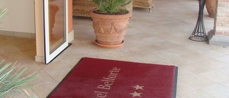 Entrada de hotel con alfombra roja, mobiliario de mimbre y una planta grande en maceta.
