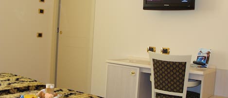 A hotel room with a bed, a desk, a chair, and a television displaying a program. There is a tray with breakfast items on the bed.